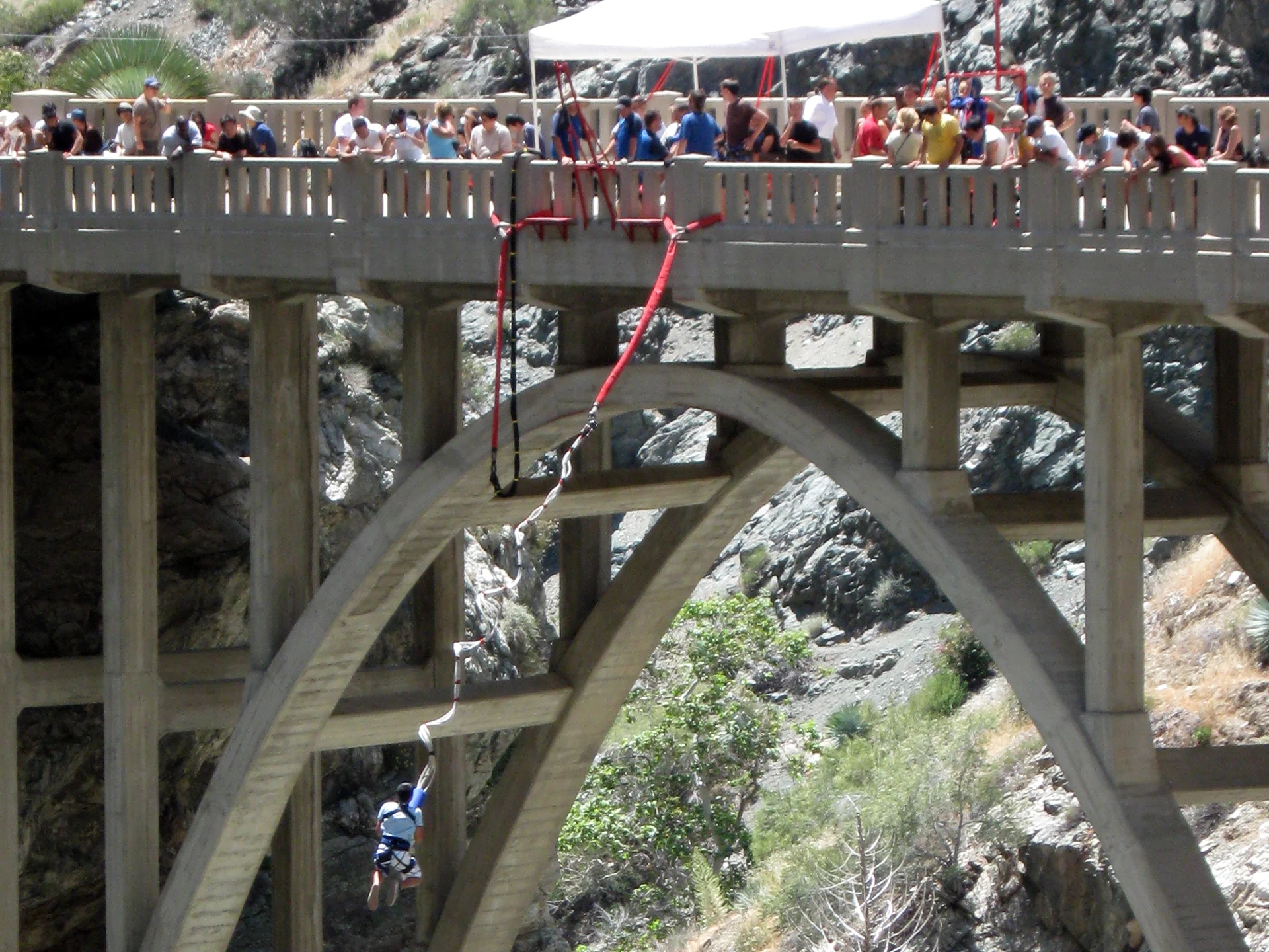 A person attached to a bungee cord below a concrete arch bridge, with spectators watching from above.