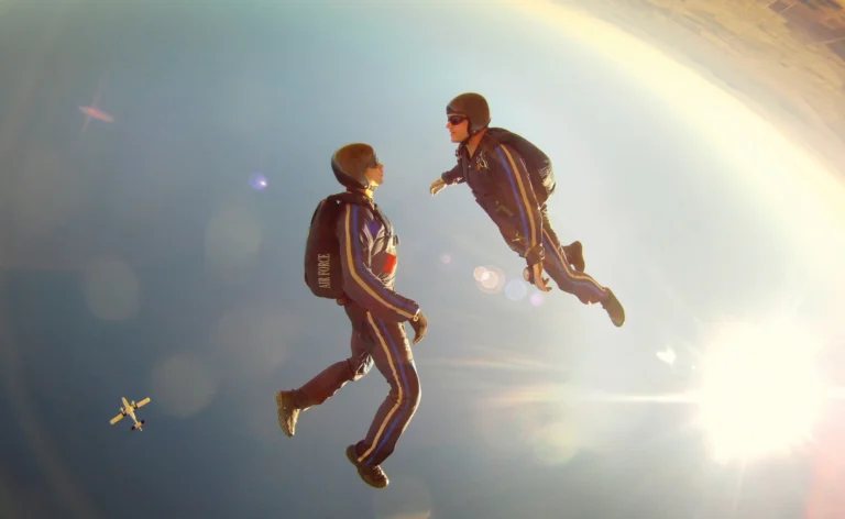 Two helmeted skydivers facing each other in freefall, with sunlight and fields visible beyond them.