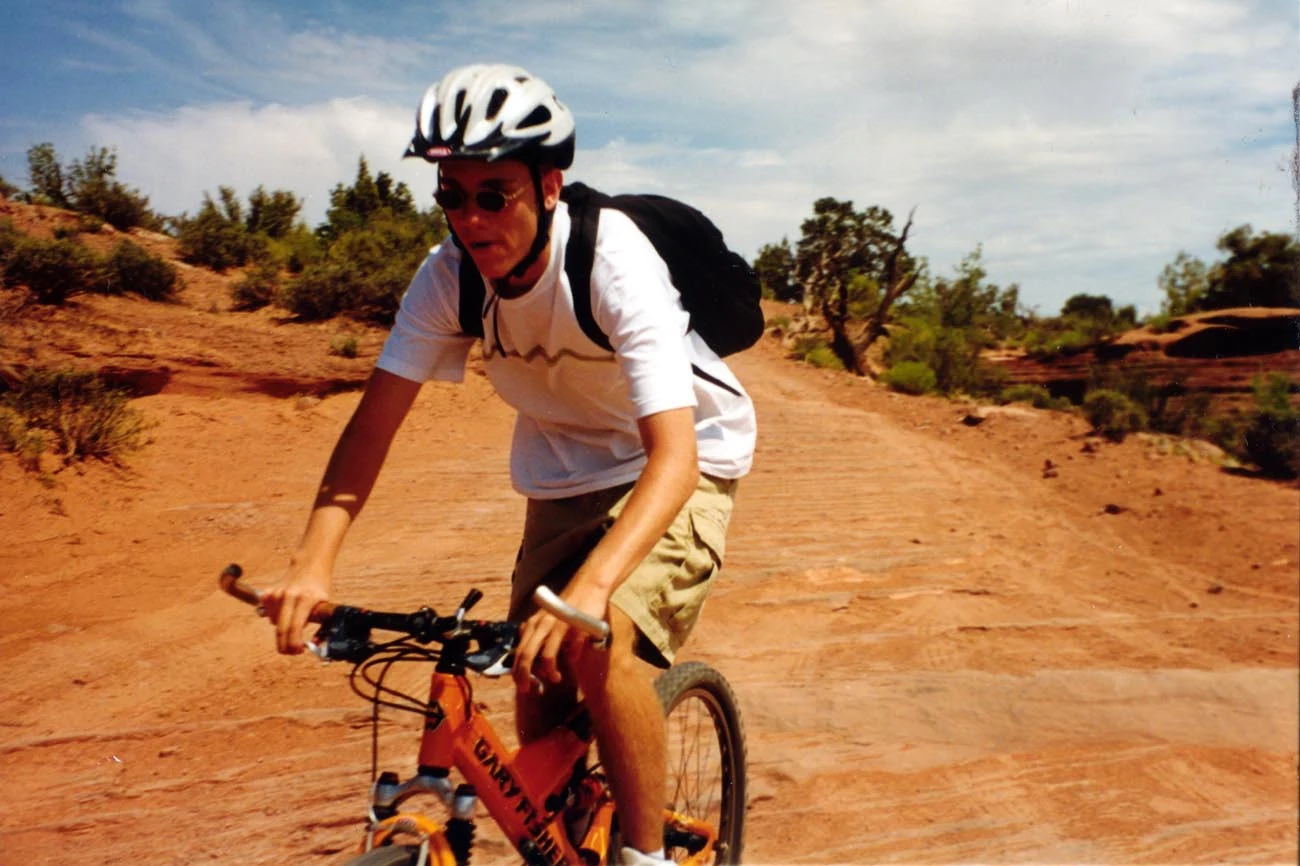 A helmeted cyclist riding a mountain bike on a broad red dirt trail, with low trees behind.