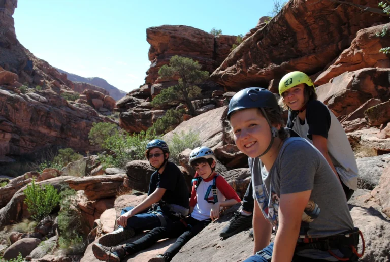 Four people wearing helmets and climbing harnesses seated beside red sandstone rocks.