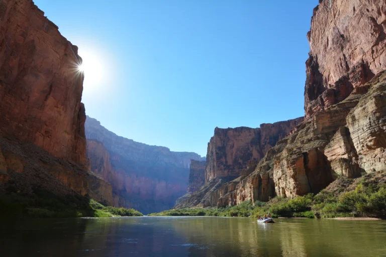 A small raft on a river beneath tall layered canyon walls, with blue sky above.