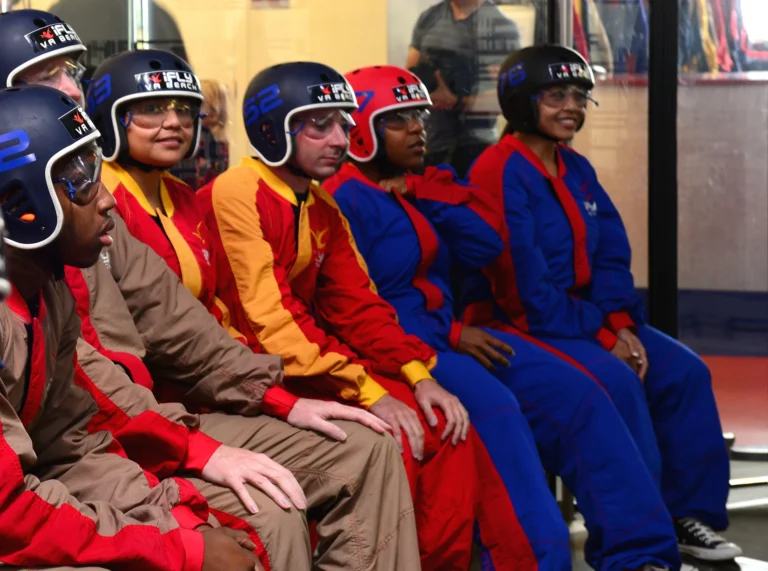 People seated indoors wearing colorful flight suits, helmets and protective goggles.