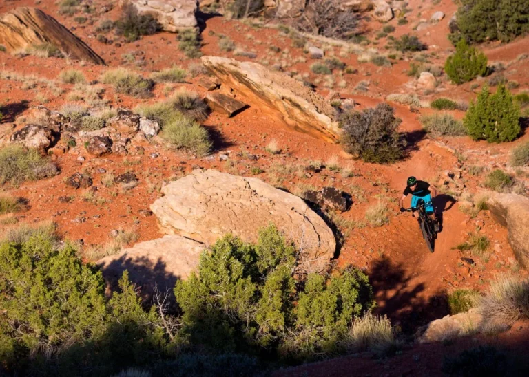 A helmeted mountain biker riding a dirt trail between red sandstone rocks and desert shrubs.