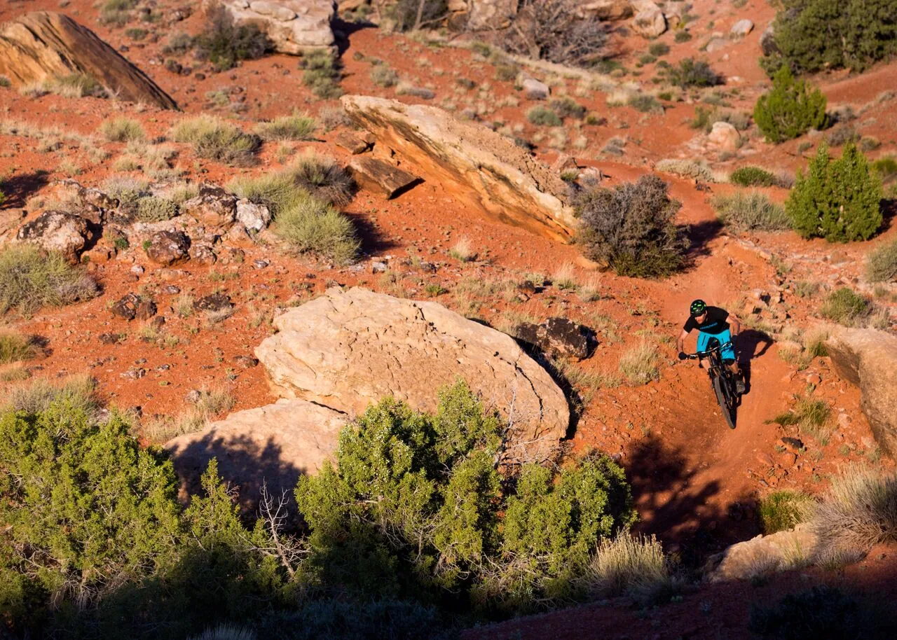 A helmeted mountain biker riding a dirt trail between red sandstone rocks and desert shrubs.