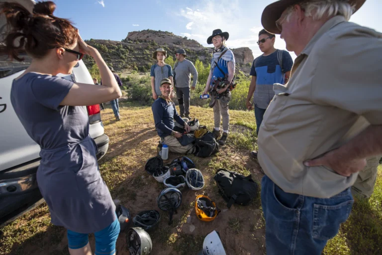 A group gathered outdoors beside helmets and climbing equipment, with rocky hills behind them.