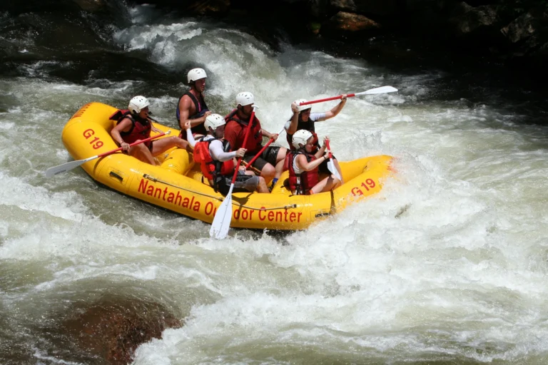 Six people wearing helmets and flotation vests in a yellow raft passing through whitewater.