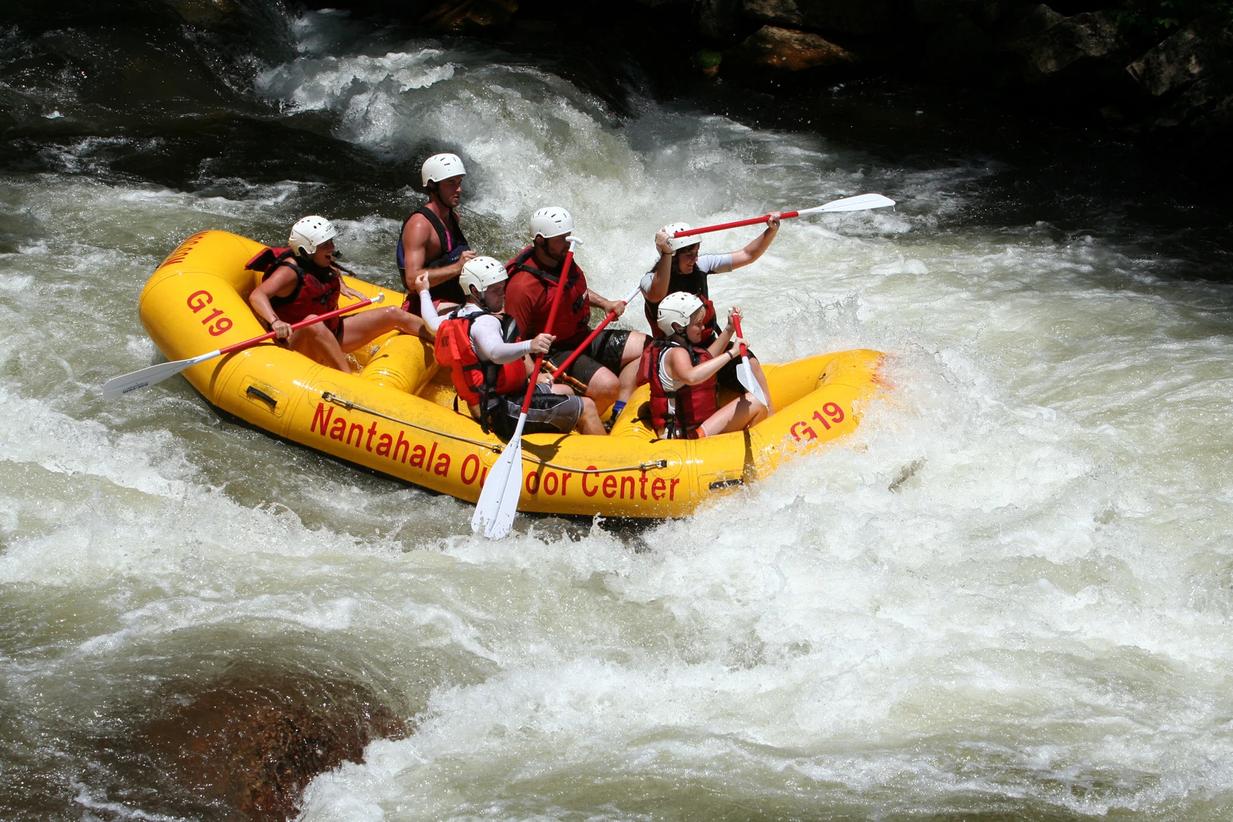 Six people wearing helmets and flotation vests in a yellow raft passing through whitewater.