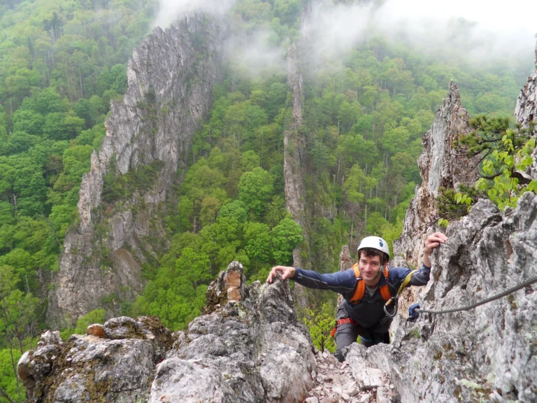 A helmeted person wearing a harness between rocks beside a fixed cable, with tall rock fins and forest behind.