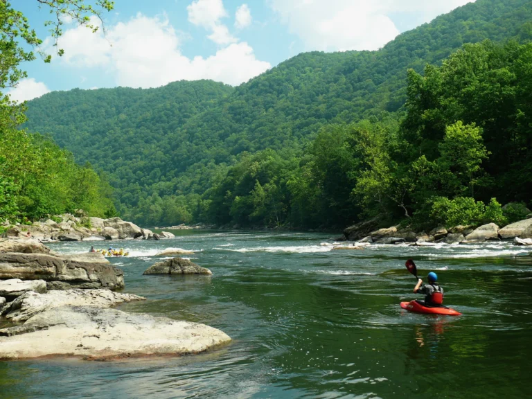 A kayaker on a river bordered by rocks and forested hills, with a raft farther upstream.