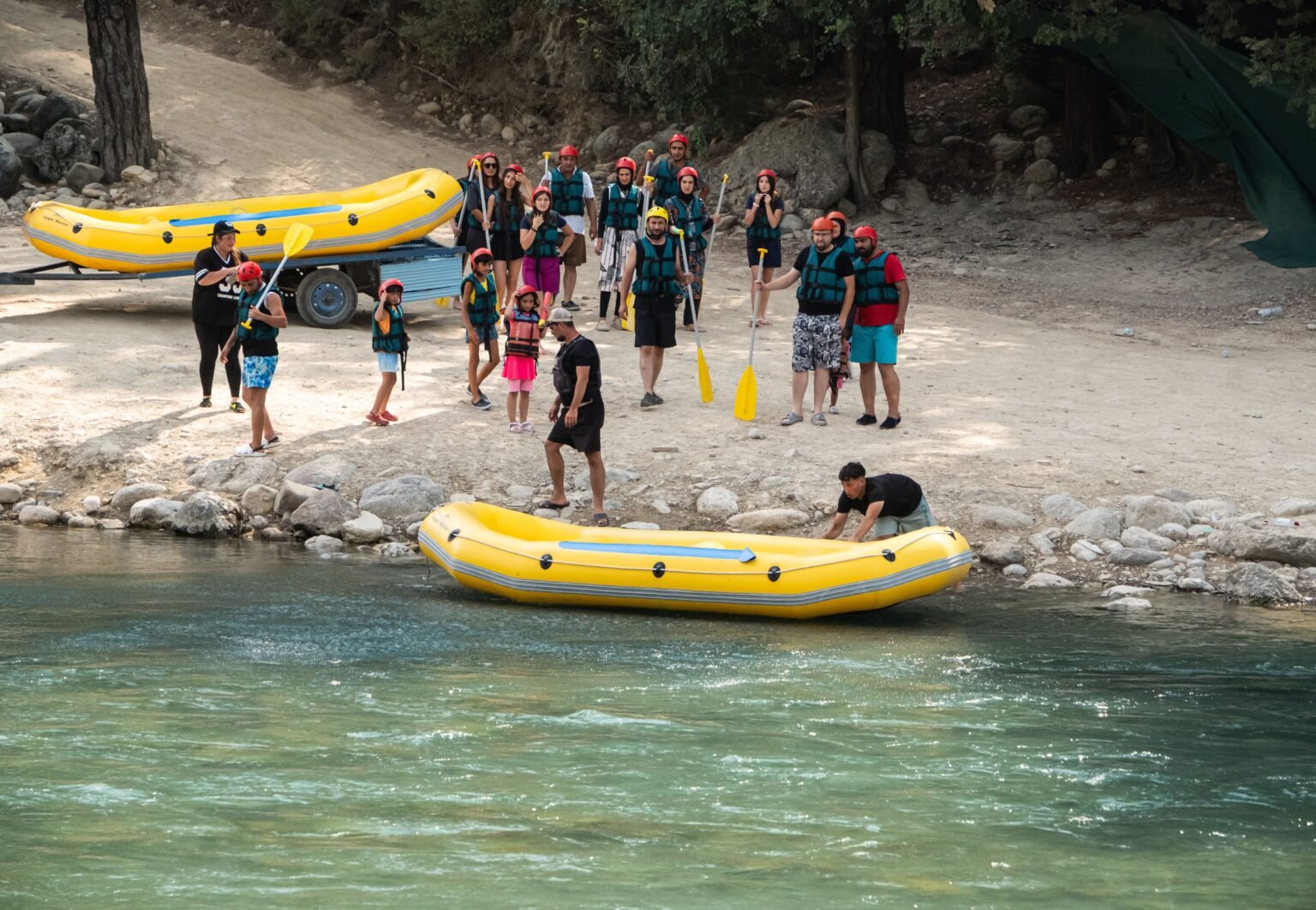 Adults and children wearing helmets and life jackets hold paddles beside yellow rafts on a riverbank.