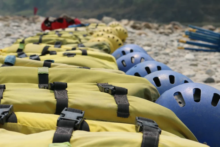 Rows of yellow flotation vests with black straps and blue and yellow helmets laid out on a rocky riverbank.