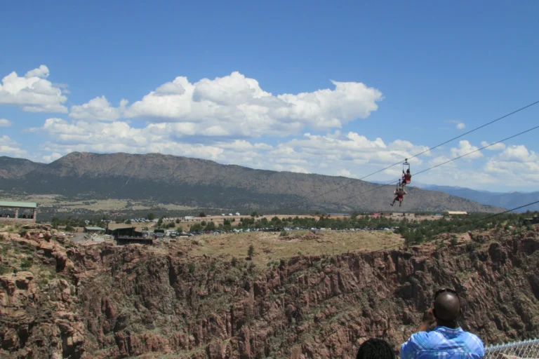 Two people suspended from parallel zipline cables above a rocky gorge, with an observer in the foreground.