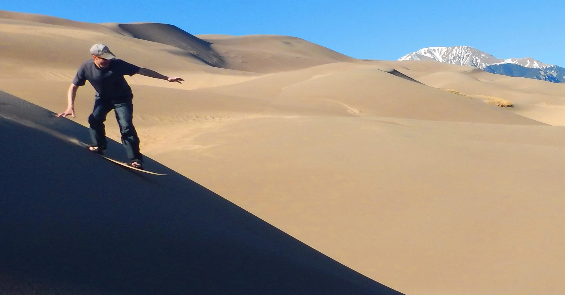 A person balancing on a sandboard on a sloping dune, with dunes and snow-covered mountains behind.