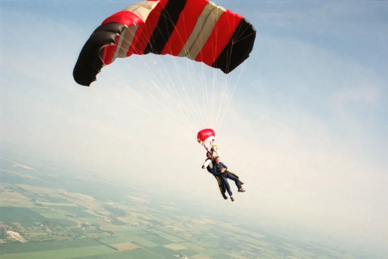 Two people suspended beneath an open black, red and white parachute, with fields visible far below.