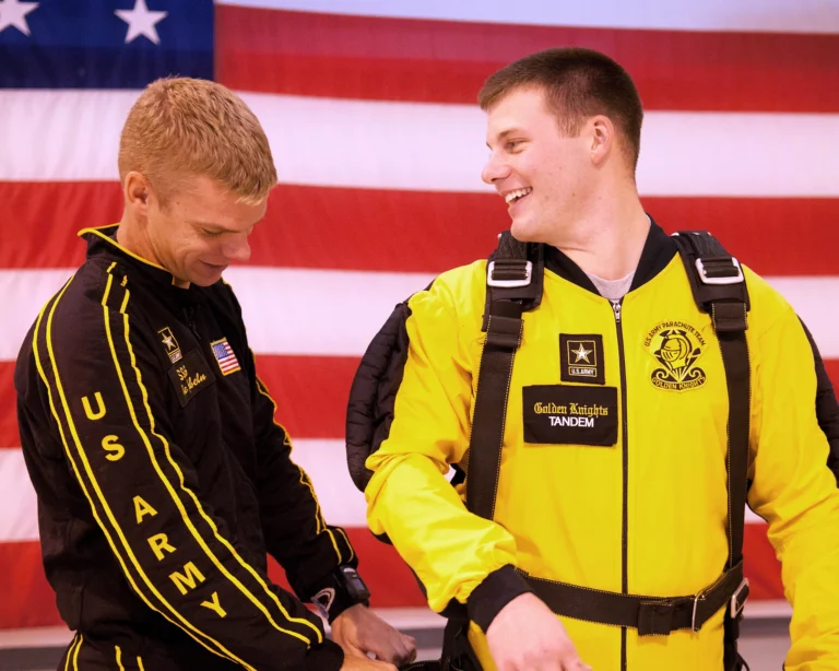 One person adjusting the harness of another person in a yellow jumpsuit, both in front of a U.S. flag.