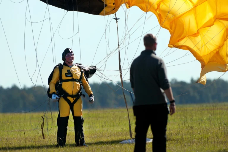 A smiling person in a yellow jumpsuit and harness beneath a partly collapsed parachute on grass, with another person in the foreground.