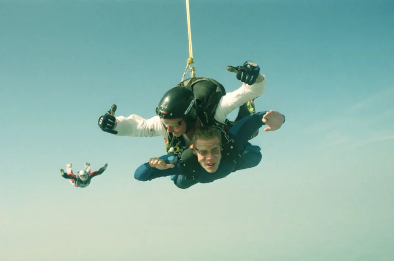 Two people attached in tandem in freefall, with another skydiver behind them against a pale blue sky.