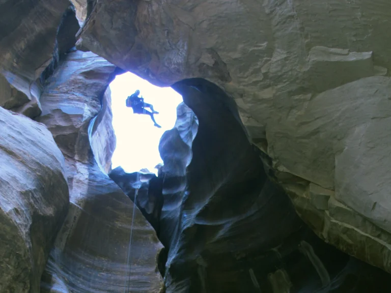 A person suspended on ropes in the bright opening between tall curved canyon walls.